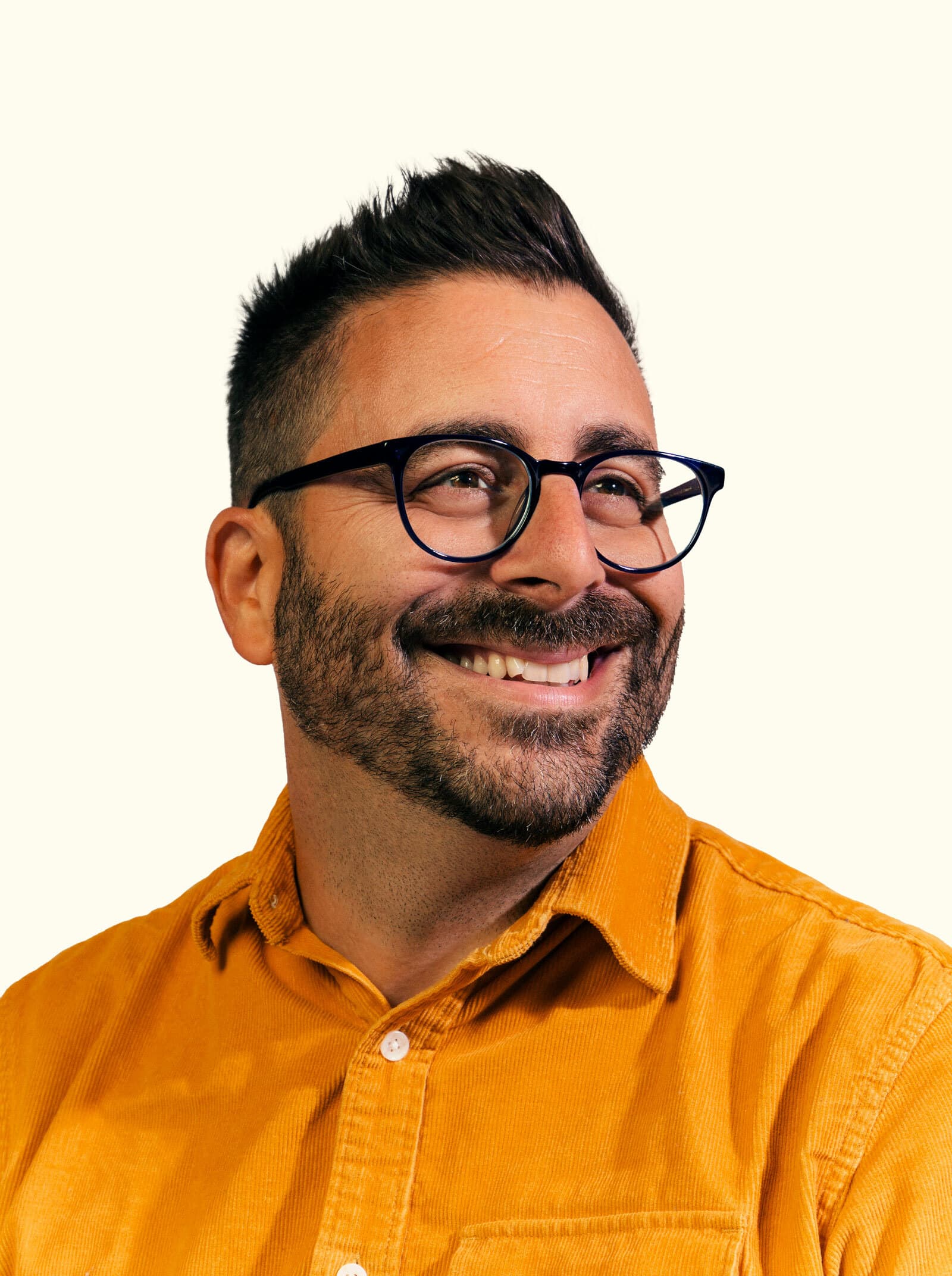 A person with short dark hair and a beard is smiling while looking to the side. They are wearing glasses and a mustard yellow button-up shirt. The background is a plain light color.