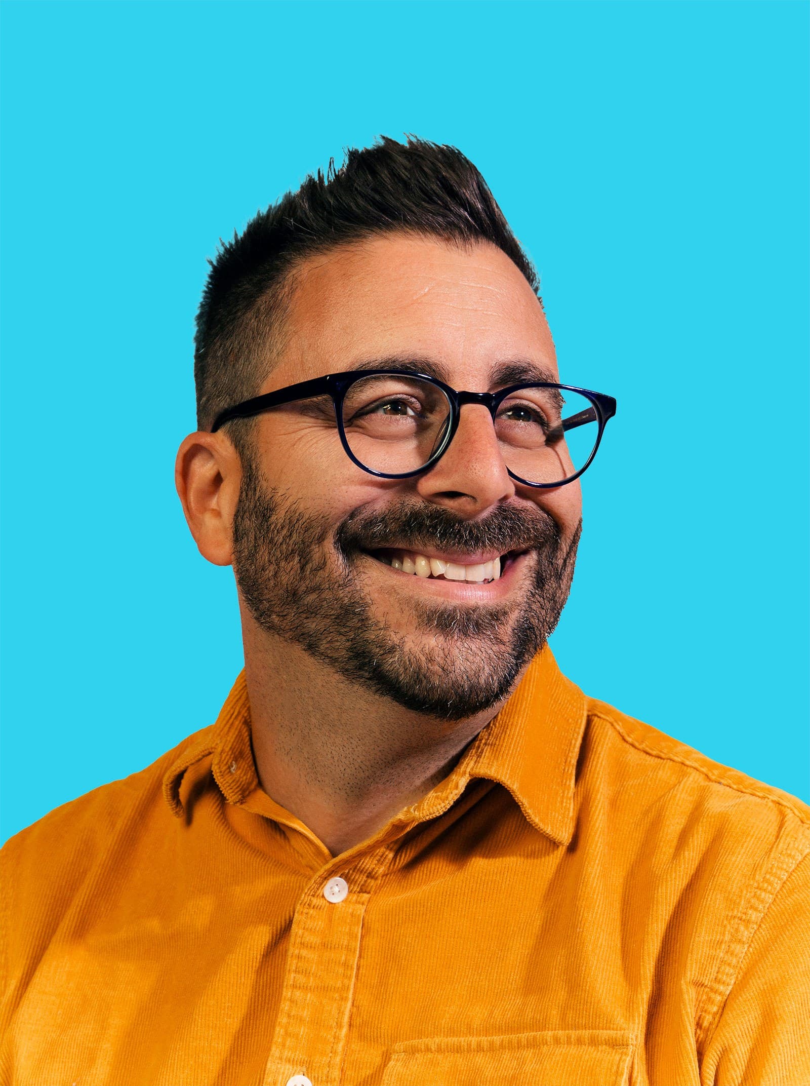 A smiling man with short dark hair, glasses, and a beard, wearing a mustard yellow shirt, against a bright blue background.