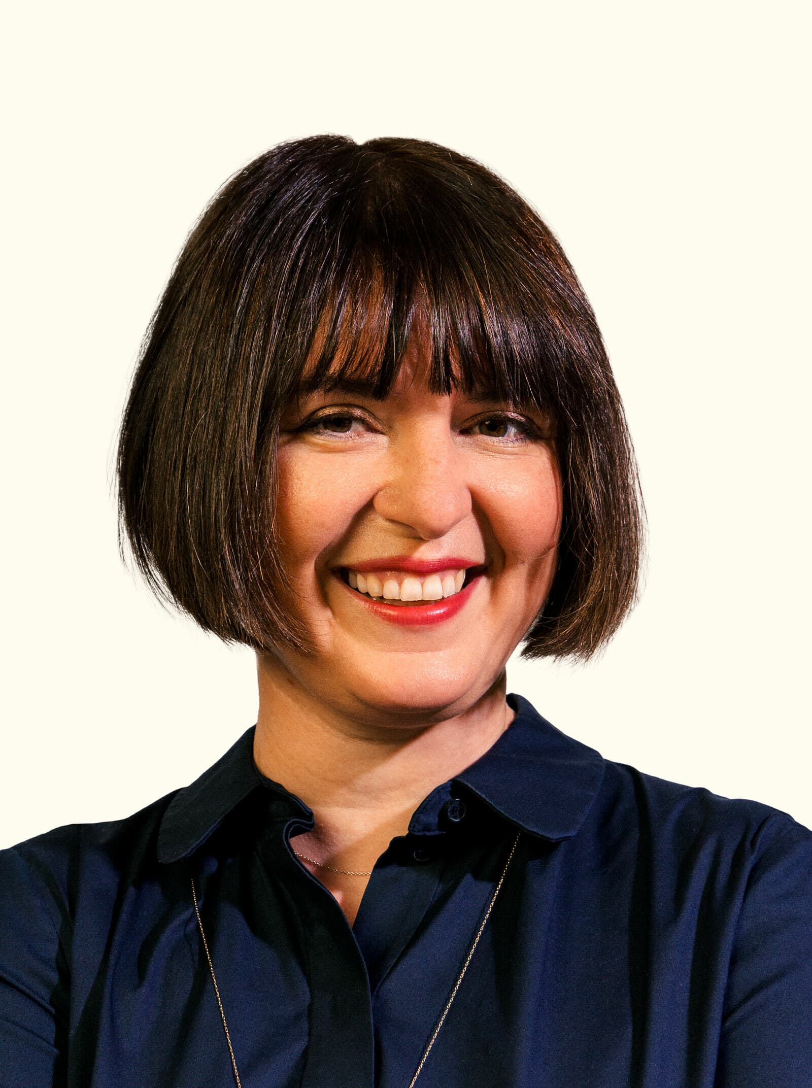 A person with short brown hair and bangs is smiling at the camera. They are wearing a dark-colored, button-up shirt and a thin, long necklace. The background is plain and light-colored.