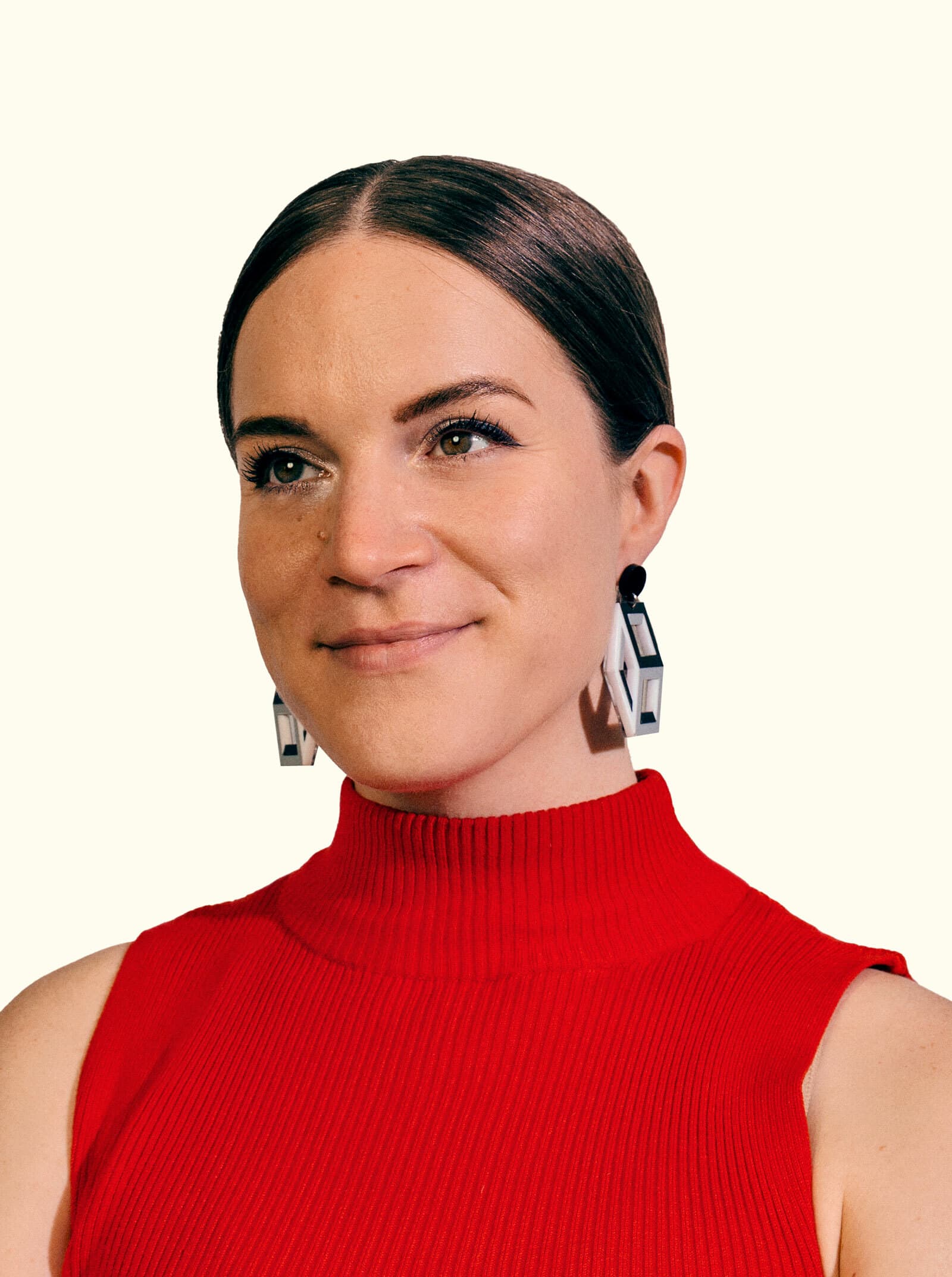 A woman with light skin and brown hair parted in the middle, wearing a red sleeveless turtleneck top and black and white geometric earrings, smiles slightly. She is posed against a plain, light-colored background.