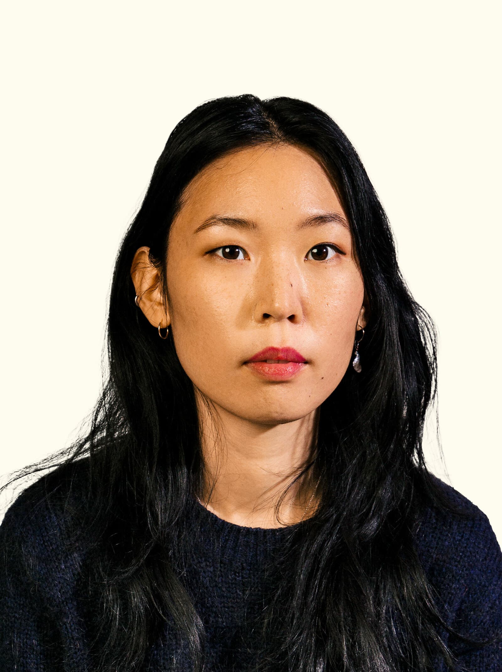 A person with long black hair, wearing a dark sweater and pearl earrings, is looking directly at the camera with a neutral expression. The background is a light, solid color.