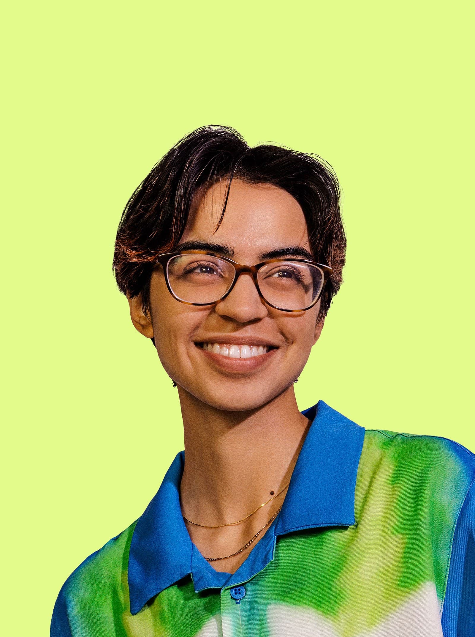 A person with short dark hair, glasses, and a bright smile wears a blue and green patterned shirt against a solid light yellow background.