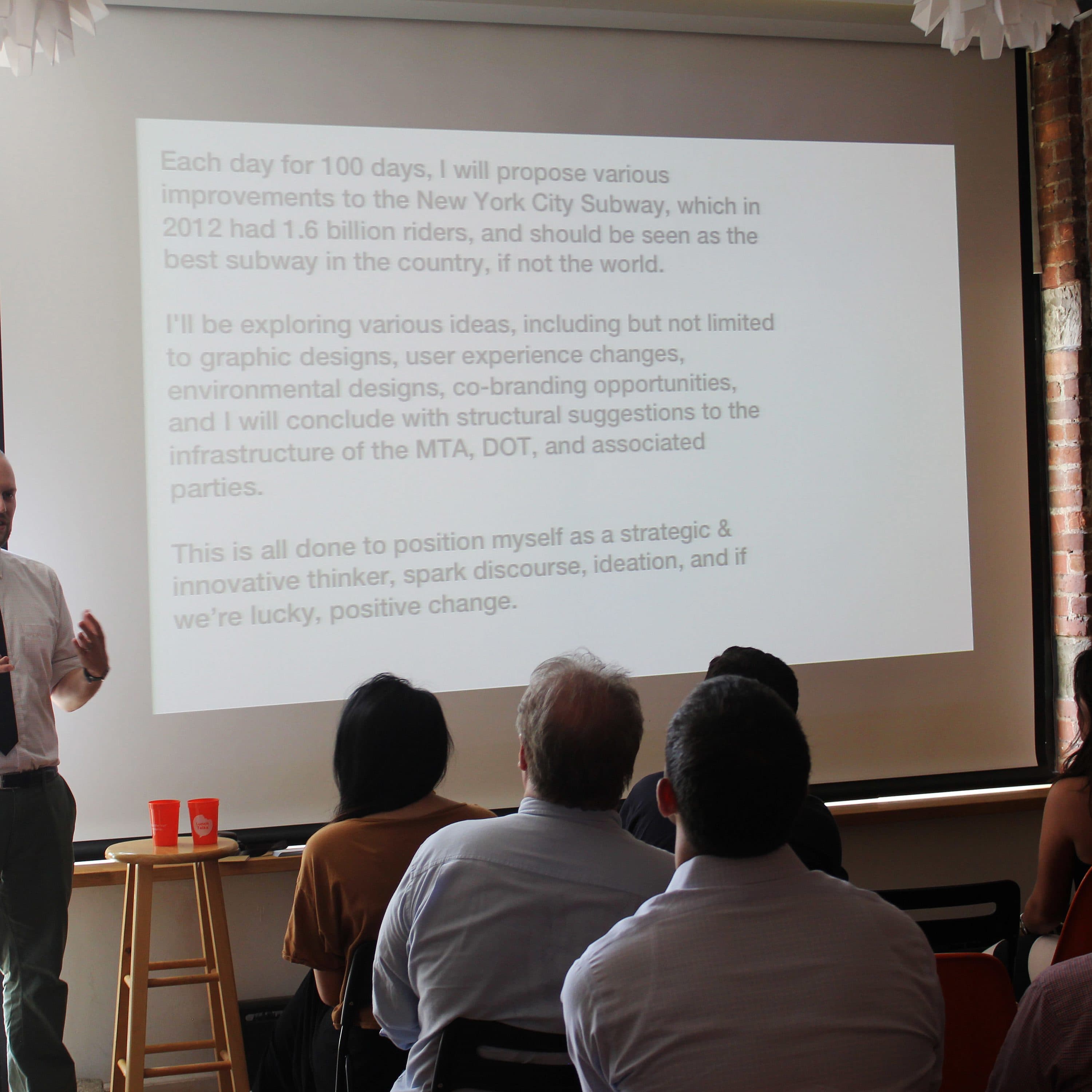 A man in a white shirt and tie stands in front of a projected presentation screen, addressing an audience seated in a room. The screen displays text about proposing improvements to the New York City Subway system over 100 days. The room has brick walls and large windows.