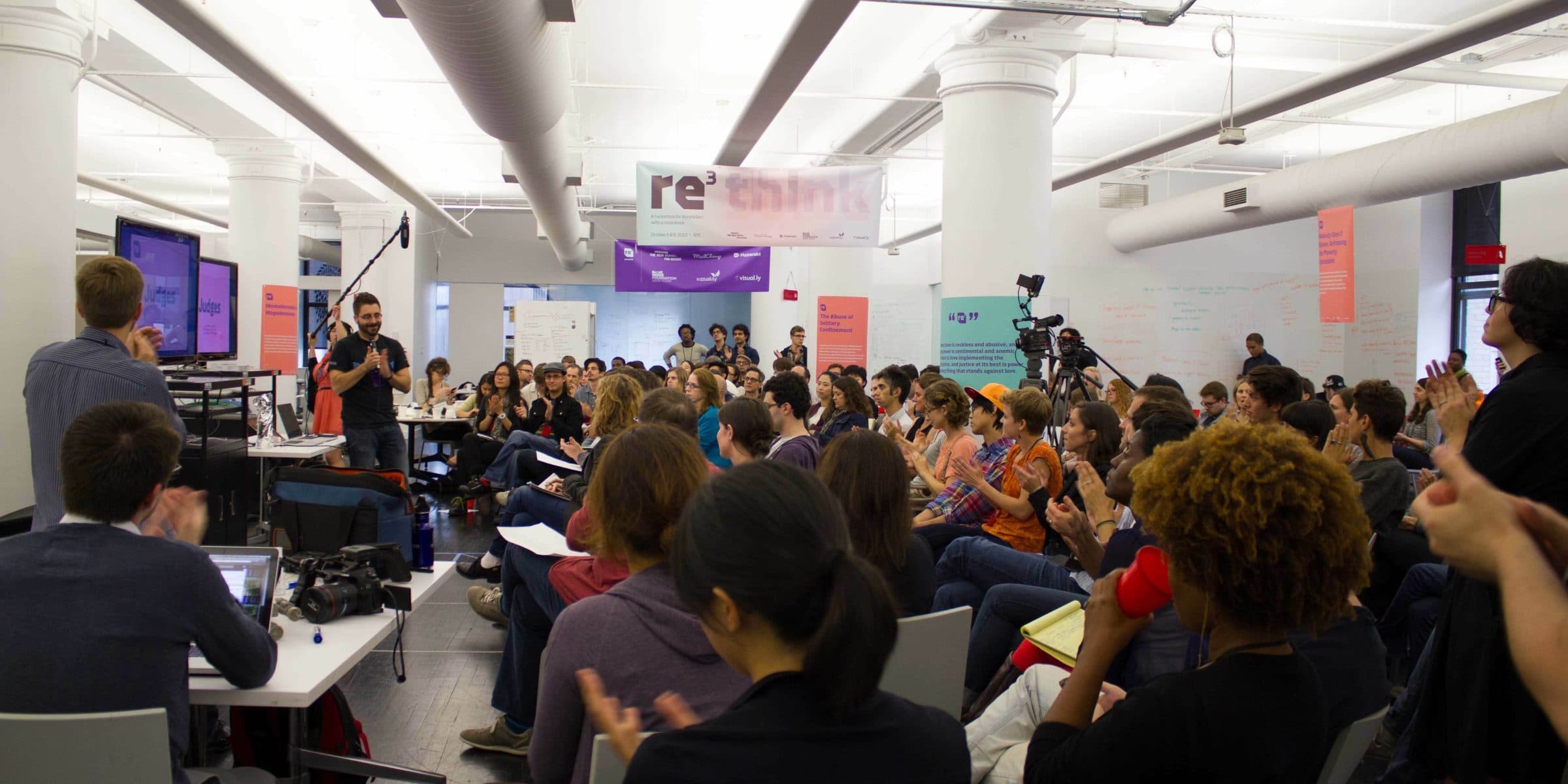 A large group of people is gathered in a modern, open-concept room with white pillars and ceiling ducts. Various individuals are engaged in conversation, taking photos, and listening attentively. A presentation screen is visible in the background, with "re:think" written above.