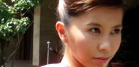 A close-up photo of a young person with light skin and dark hair pulled back in a bun. The person is outdoors, and there is lush greenery in the background. They have a serious expression and the sunlight illuminates their face.