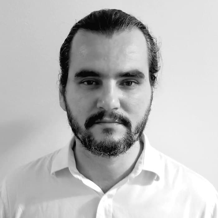 A bearded man with long hair tied back is looking directly at the camera with a neutral expression. He is wearing a white collared shirt and is positioned against a plain background. The image is in black and white.