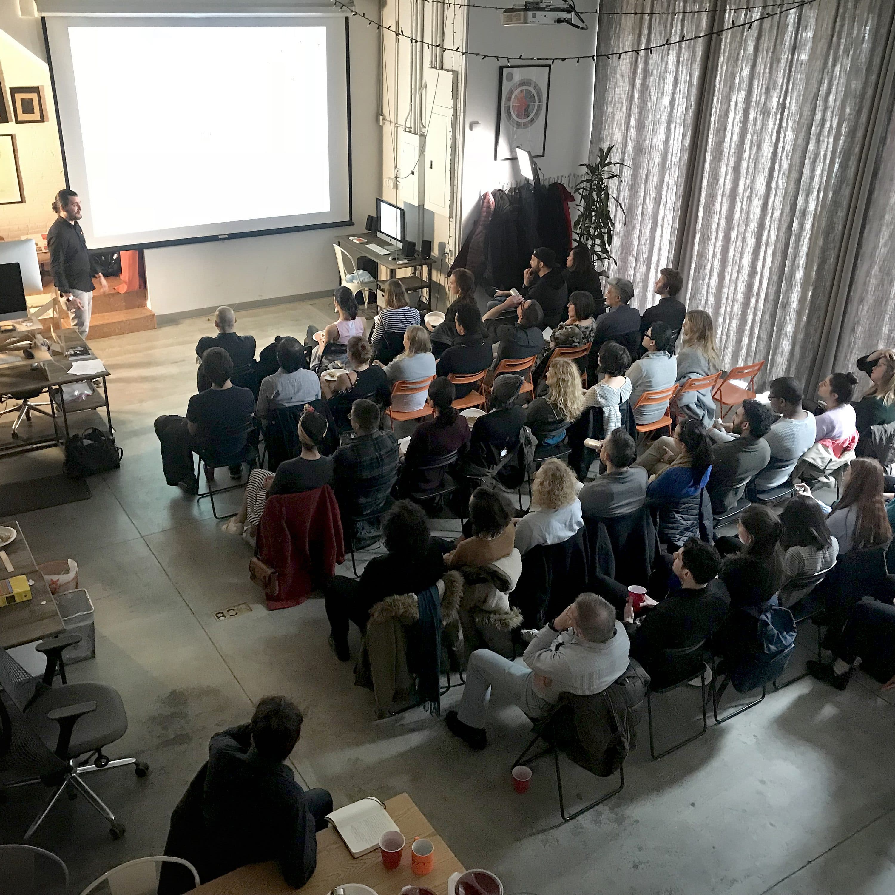 A group of people seated in a room watching a presentation on a large screen. The room has a mix of office and casual seating, including tables, chairs, and couches. The presenter is standing to the left of the screen. The room is well-lit with large windows and curtains.