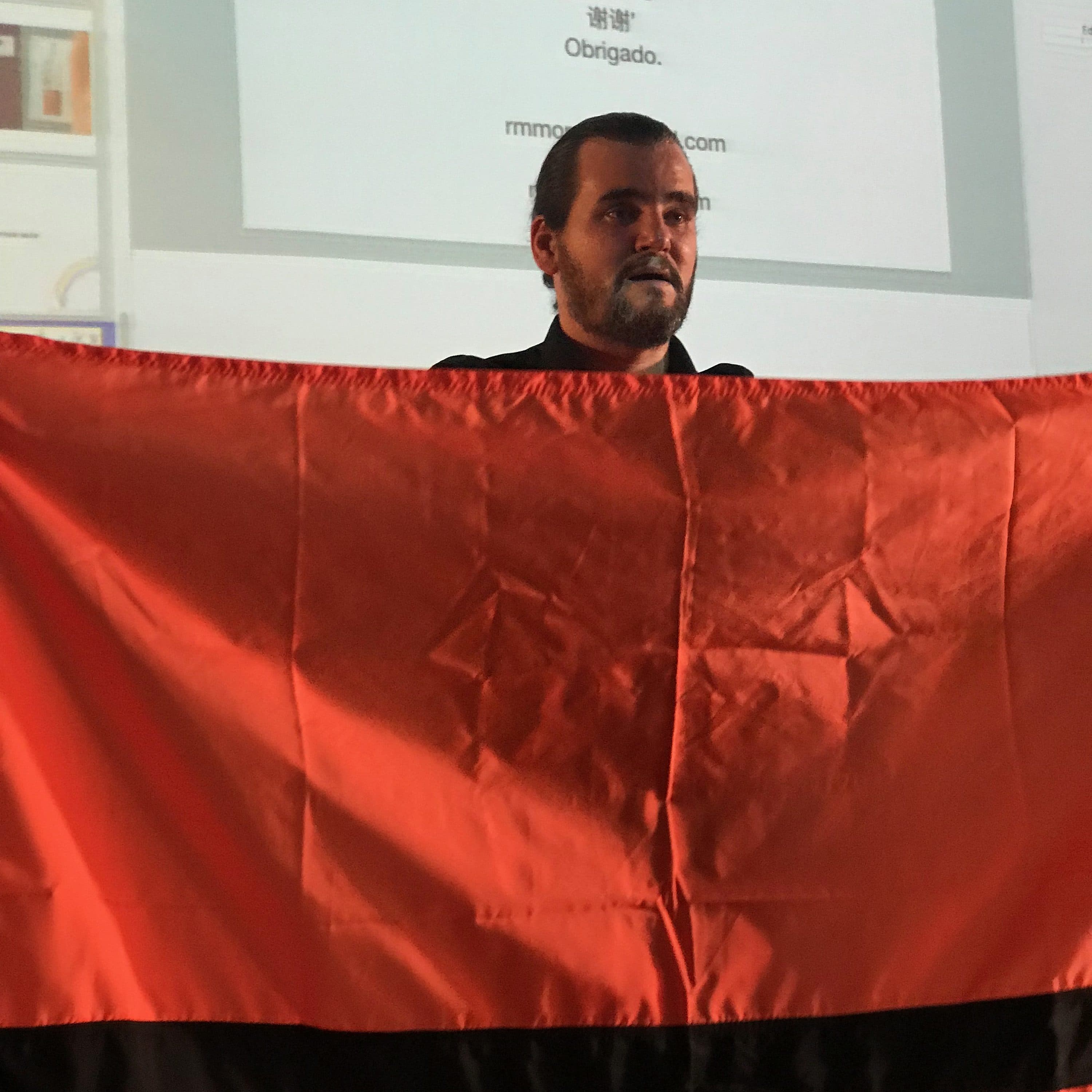 A man stands holding a large red flag with a black stripe across the middle. Behind him is a projected screen displaying a notice that reads "Pirates wanted" along with "Thank you" in multiple languages. Various text and images are also visible on the screen.