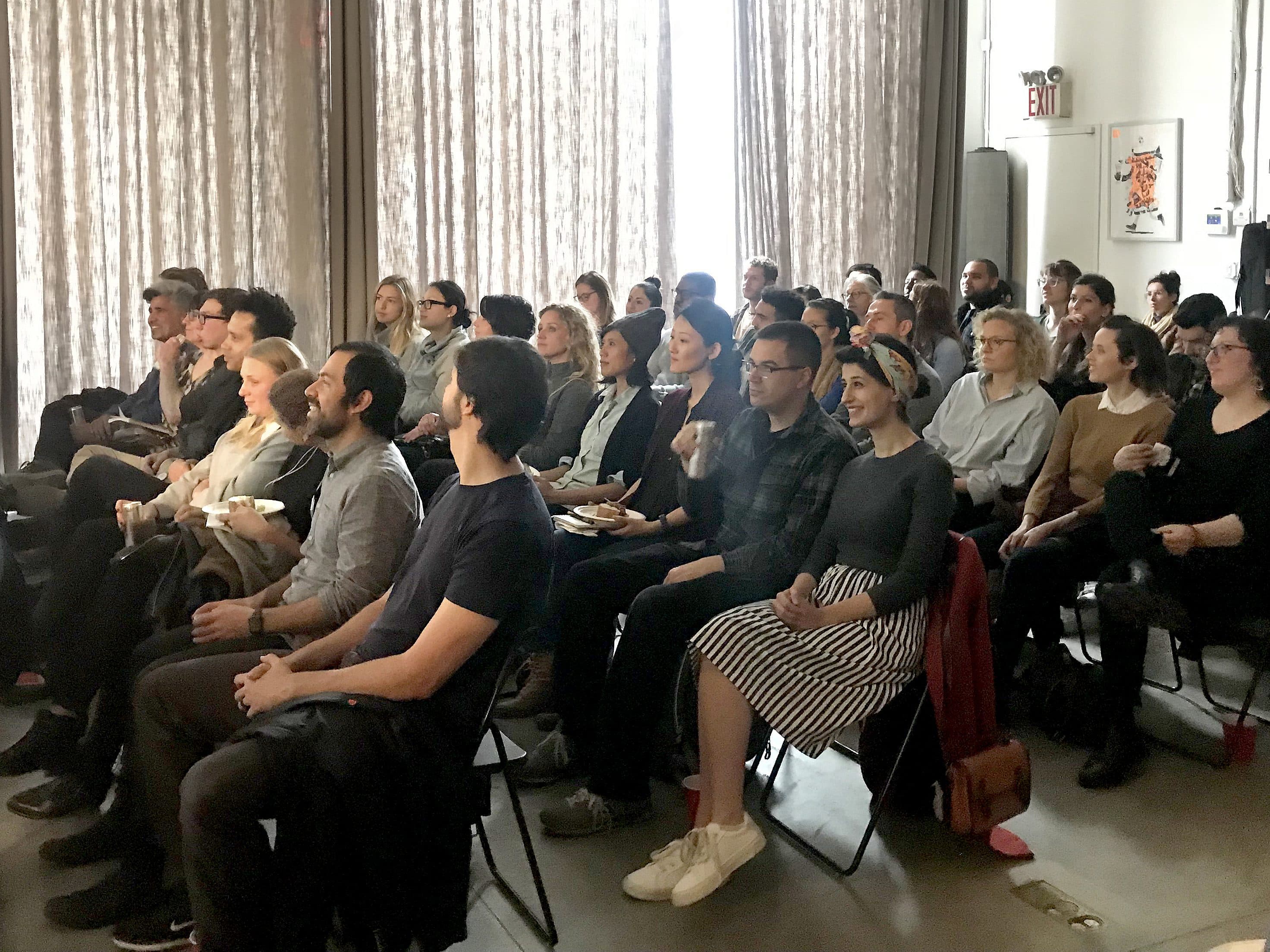 A diverse group of people sitting in rows of chairs, attentively watching a presentation in a room with large curtains. Some individuals hold notepads and drinks, while a camera setup is visible in the background. The atmosphere appears focused and engaged.