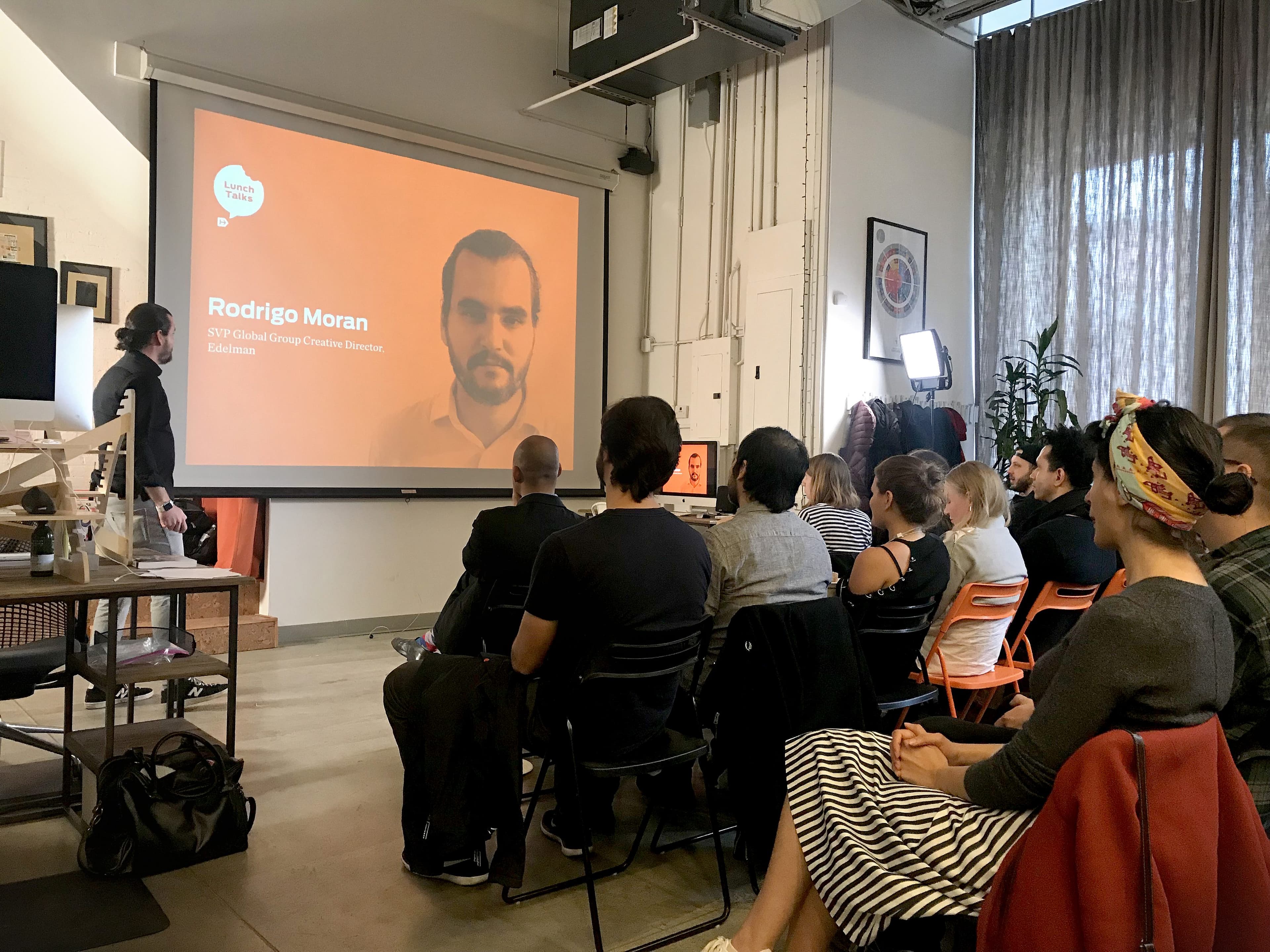 A group of people is seated and facing a large screen in a modern, brightly lit room. The screen displays the image and name of Rodrigo Moran, with his title. A speaker is standing next to the screen, addressing the audience. The room has eclectic decor and large windows.