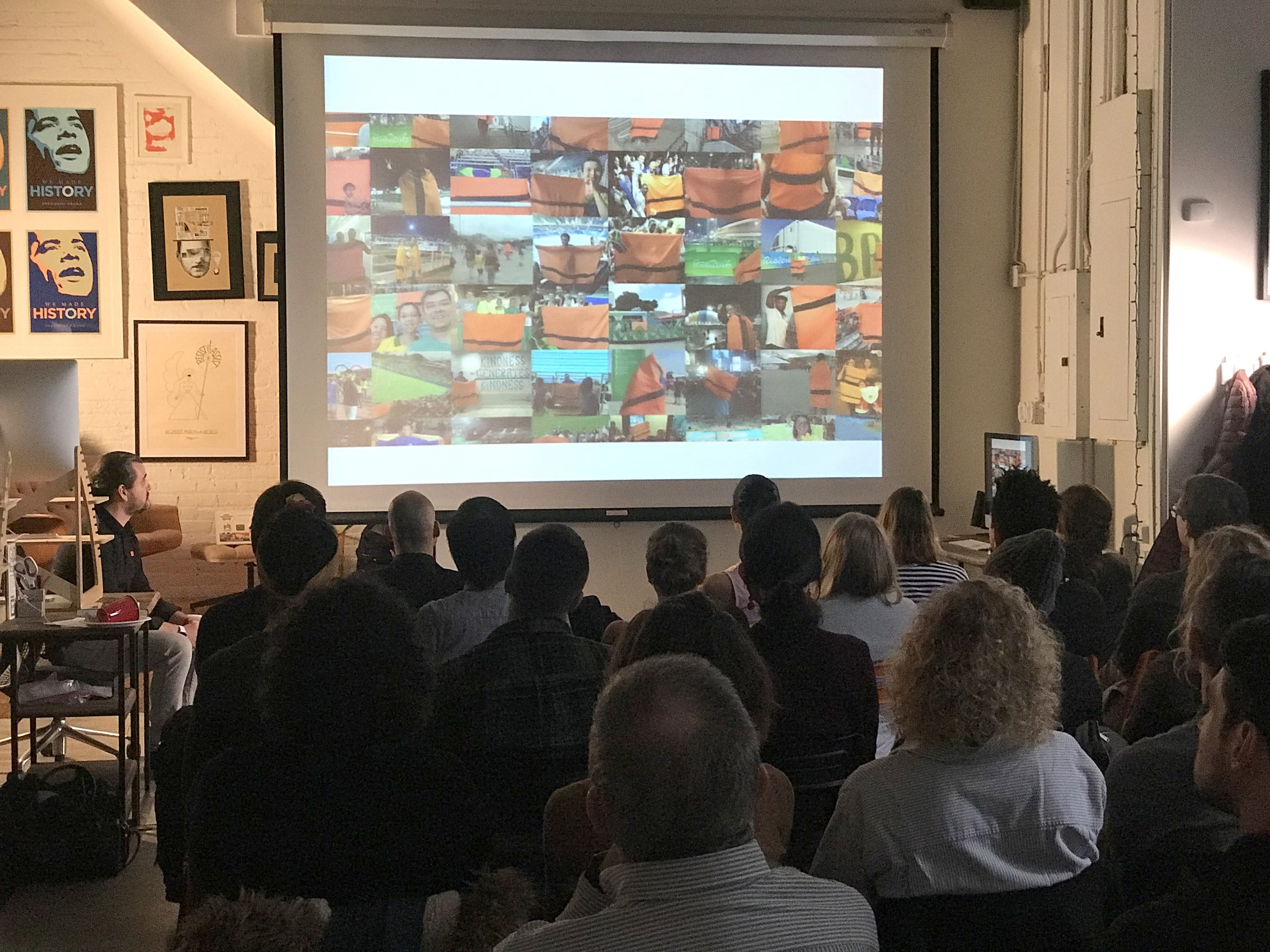 A group of people sits in a dimly lit room, watching a large screen displaying a collage of various colorful images. The audience is facing the screen, and the walls of the room have framed pictures and posters. Natural light comes in from a side window.