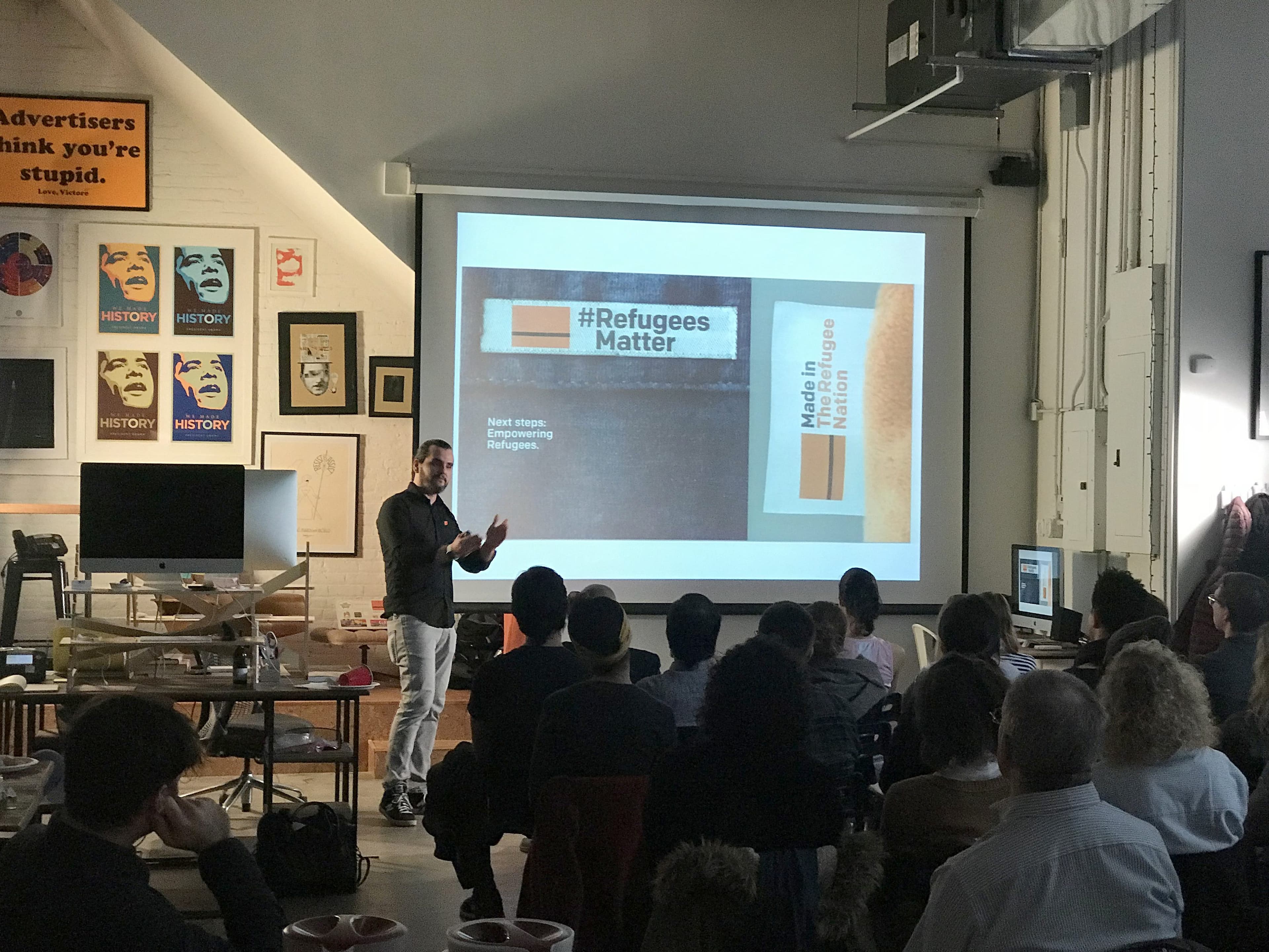 A presenter stands in front of a projected screen with the text "#RefugeesMatter" and "Made in Refugees". The audience is seated, attentively watching the presentation. The room is decorated with various posters and art on the walls.