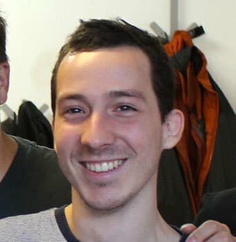A person with short dark hair and a light beard is smiling, looking directly at the camera. He is wearing a light-colored shirt. Coats and jackets are hanging in the background.