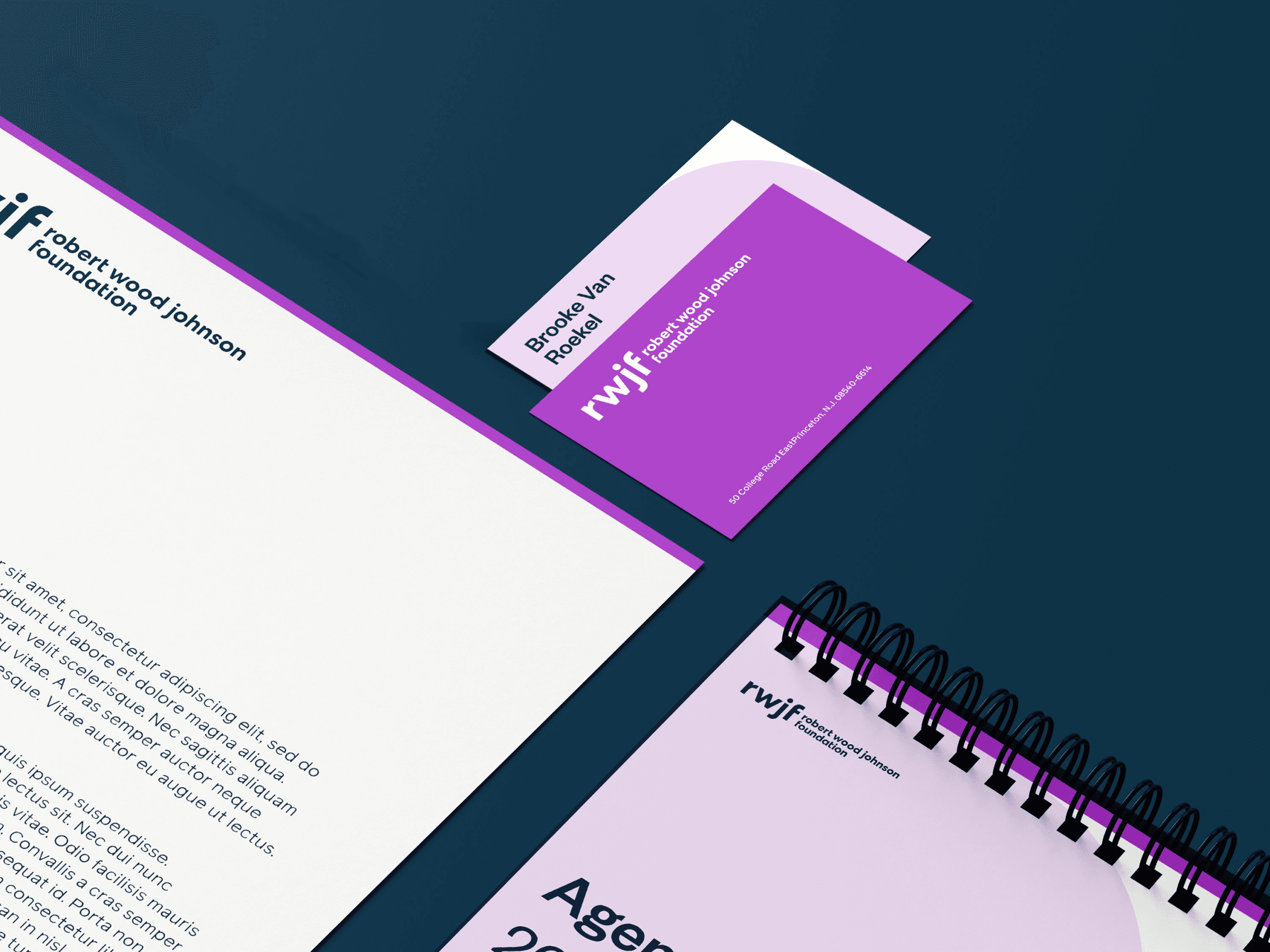 Flat lay of branded office materials including a letterhead, business card, and spiral-bound agenda. The materials feature a purple and white color scheme with text related to the Robert Wood Johnson Foundation. The items are arranged on a dark blue surface.