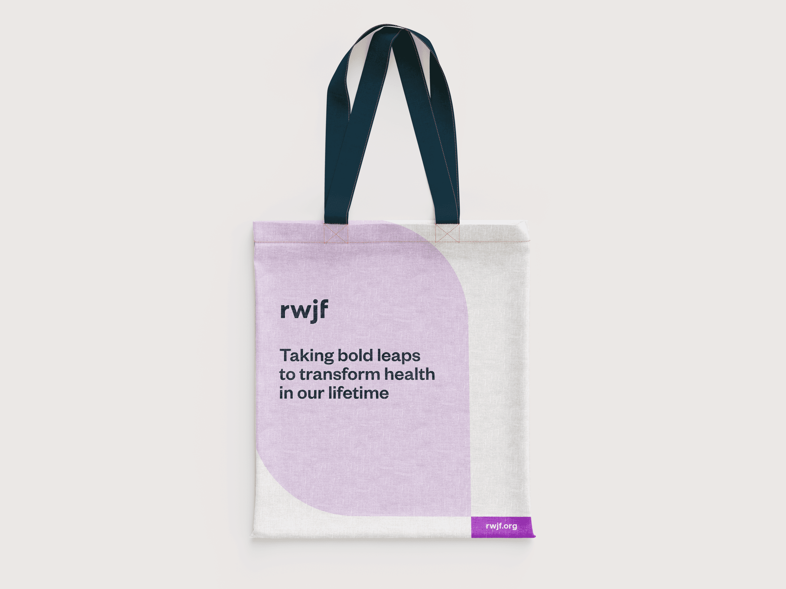 A white tote bag with dark blue handles features the text "rwjf" and "Taking bold leaps to transform health in our lifetime." The bag has a purple semicircle design in the bottom right corner and the website "rwjf.org" in white text on a small purple rectangle.