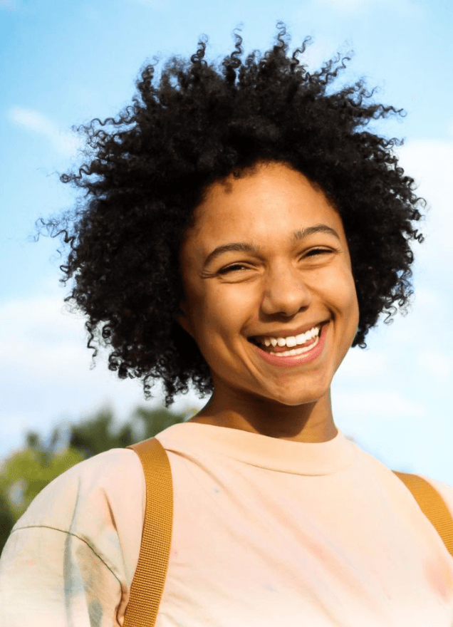 A person with curly hair smiles warmly at the camera. They are wearing a light-colored shirt and have a yellow-brown strap on their shoulder. The background shows a clear sky with some clouds and trees in the distance.