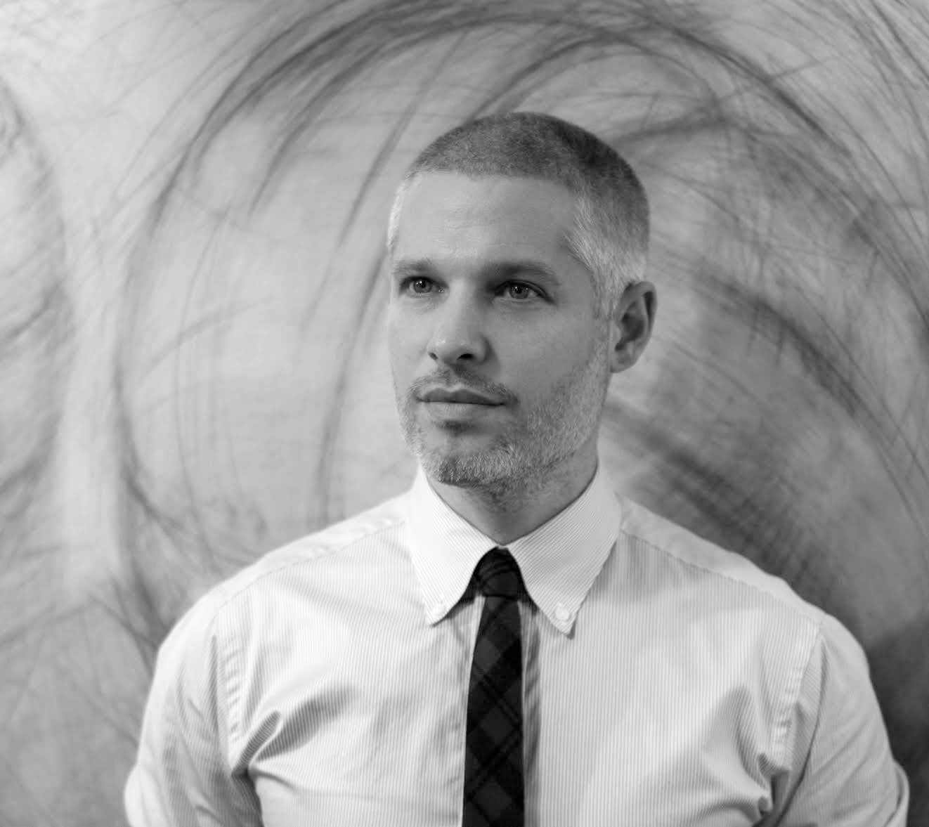 A grayscale photo of a man with short-cropped hair and a beard, wearing a light-colored dress shirt and a dark plaid tie, standing in front of an abstract background with circular, swirling patterns. He is gazing slightly to the side with a focused expression.