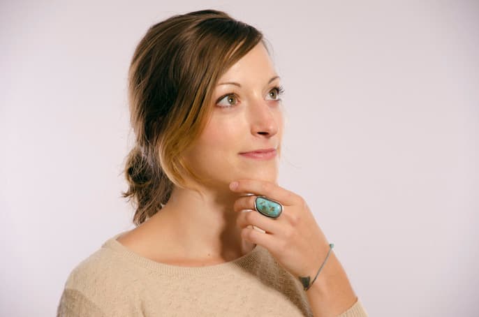 A woman with light brown hair tied back, wearing a beige sweater, looks thoughtfully to the side. She rests her chin on her hand, revealing a large turquoise ring on her finger. The background is a plain light gray.