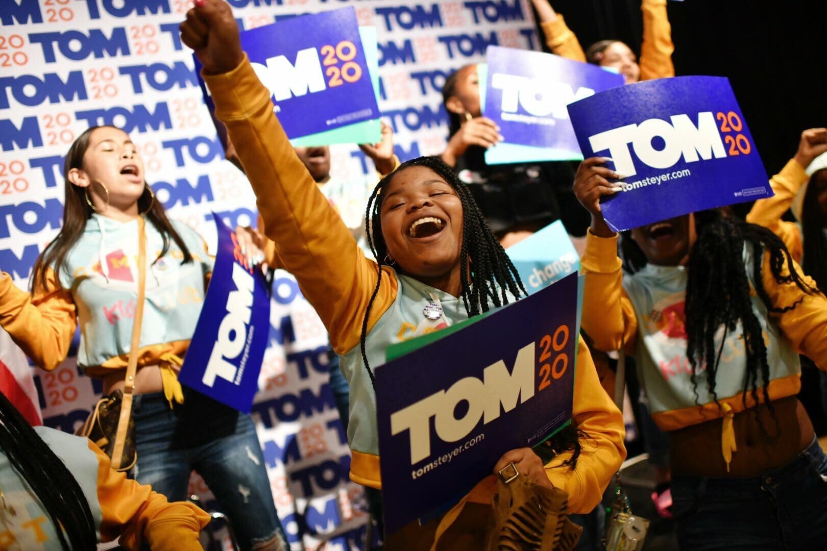 A group of enthusiastic supporters cheers and raises signs that read "TOM 2020" at a political rally. They are wearing matching outfits and expressing excitement. The background is filled with more "TOM 2020" banners.