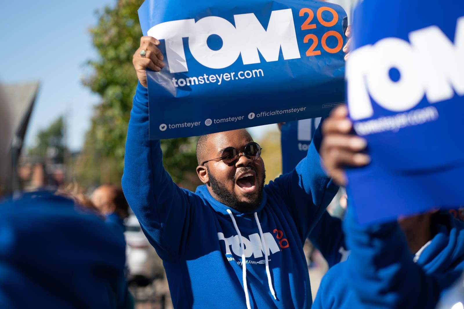 A man wearing sunglasses and a blue hoodie with "TOM 2020" is holding up a "TOM 2020" campaign sign. He is surrounded by others holding identical signs in a rally setting. Trees and sunlight in the background suggest an outdoor event.