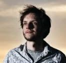 A person with light skin and medium-length dark hair stands outside, looking slightly to the side. They are wearing a light-colored, patterned shirt and a gray t-shirt underneath. The sky in the background is softly lit with a warm, golden hue.