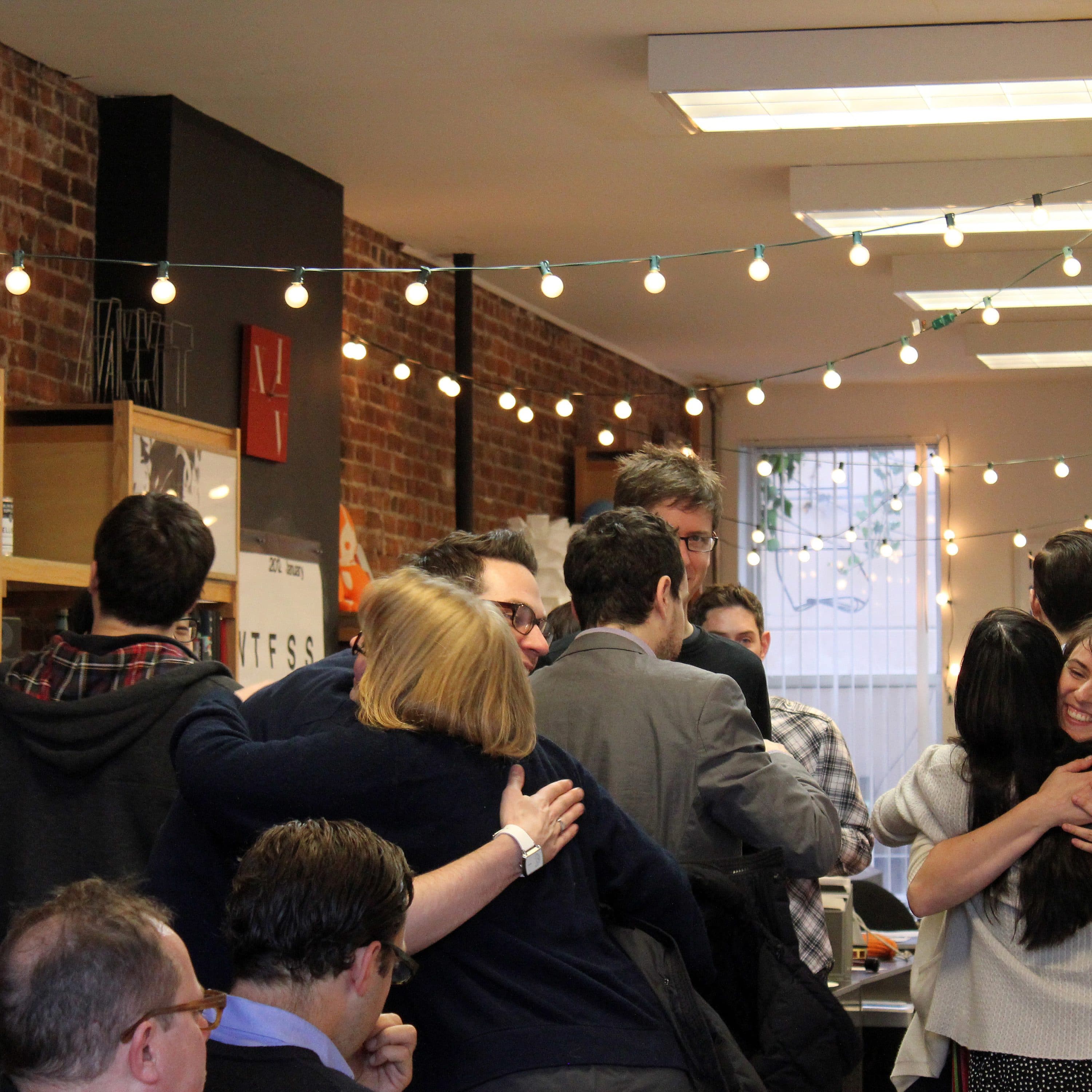 A group of adults congregate in a warmly lit room adorned with string lights and brick walls. People are hugging and engaging in conversations, creating a friendly, social atmosphere. Shelves with books and decorations are visible in the background.