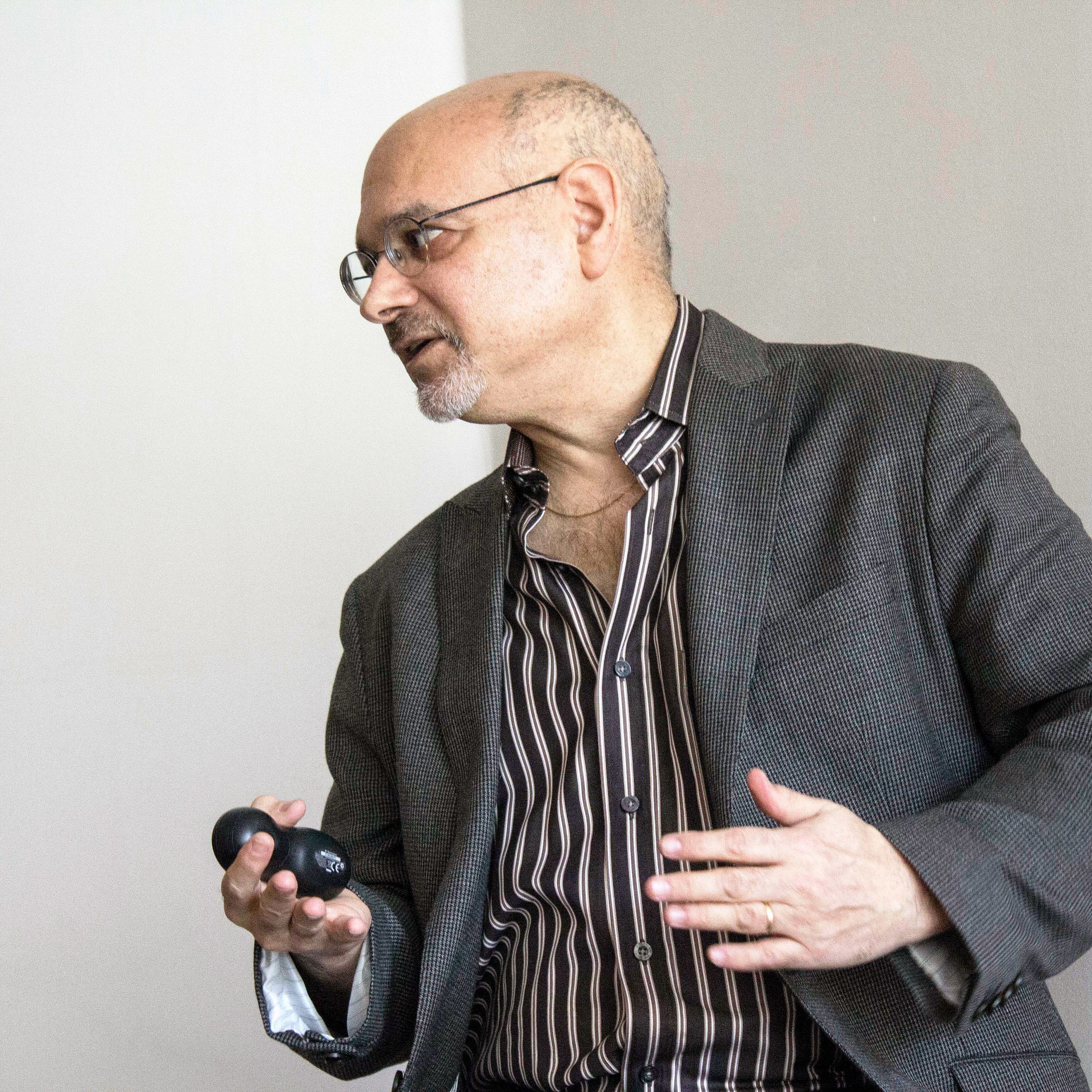 A man with glasses, a beard, and a bald head is speaking. He is dressed in a striped shirt under a gray blazer and is holding an open black eyeglass case. His right hand is gesturing while he speaks against a neutral background.