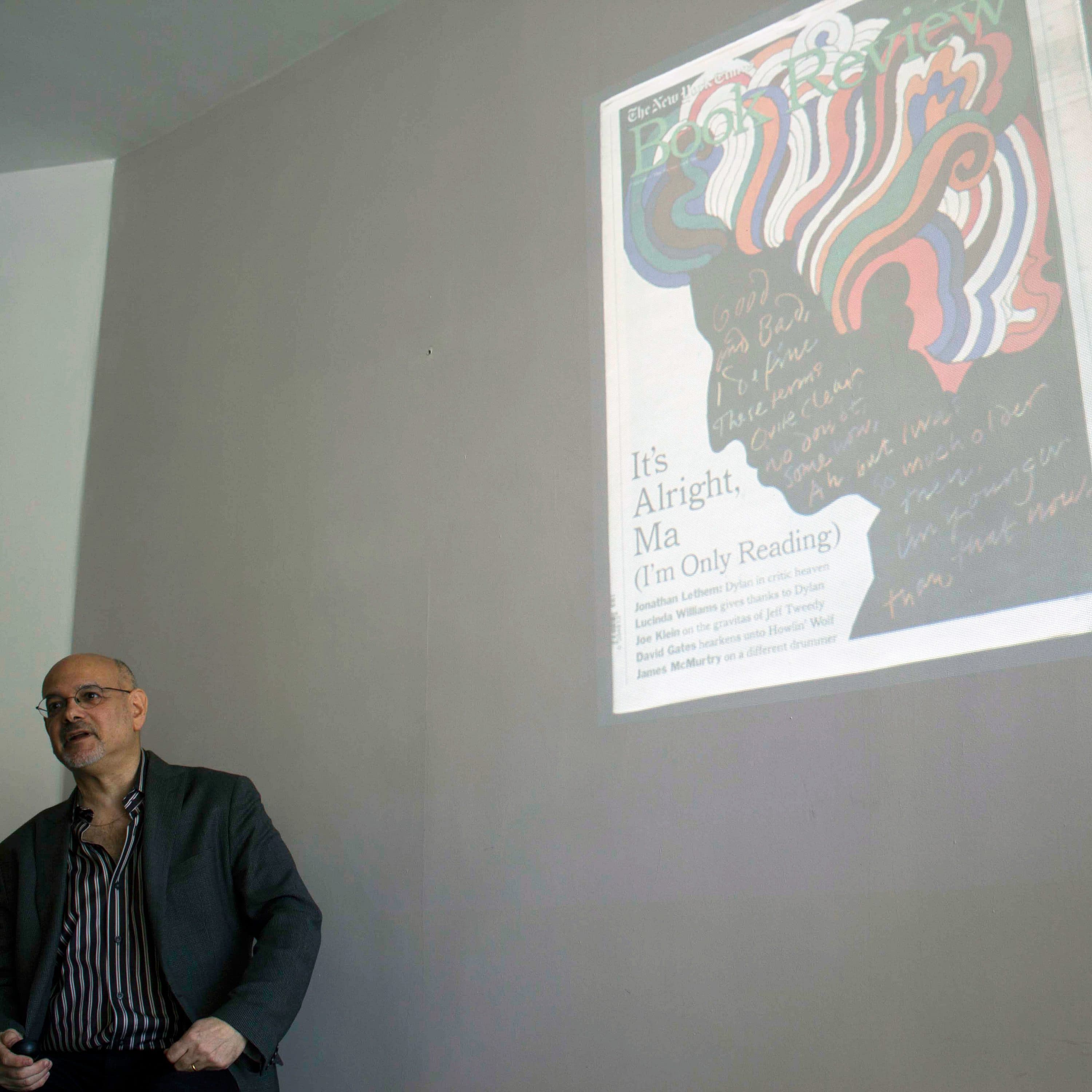 A person is standing next to a wall with a magazine cover projected onto it. The projection features colorful artwork and text that reads "It's Alright, Ma (I'm Only Reading)." The person is dressed in a dark jacket and has a serious expression.