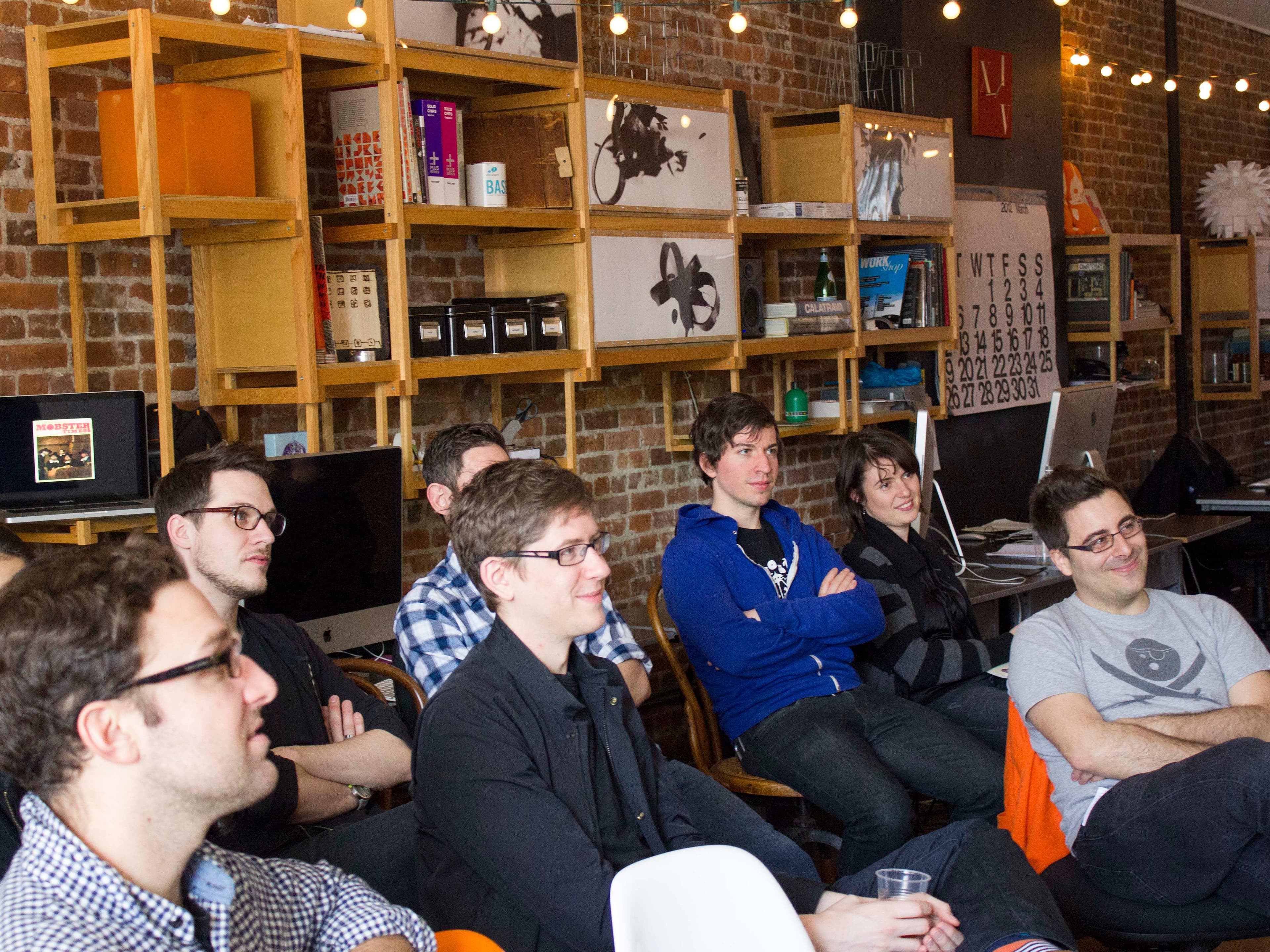A group of people are seated and listening attentively in a casual office setting. Shelving on a brick wall behind them holds books, decorations, and electronics. String lights are hung across the room, adding a warm ambiance to the scene.