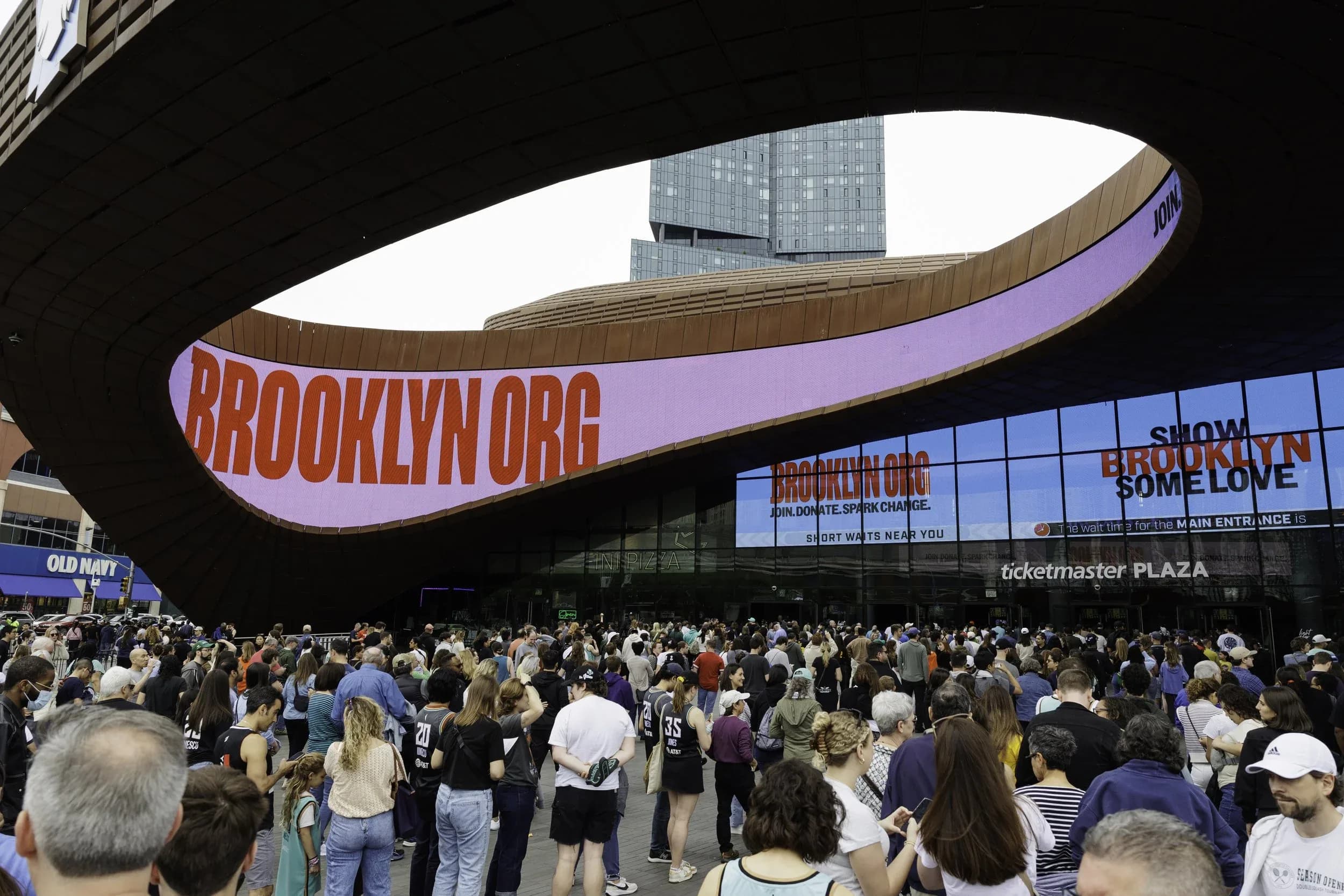 A large crowd gathers outside the Barclays Center in Brooklyn, New York, beneath a curved digital marquee displaying “BROOKLYN.ORG” and other messages. The scene is lively with people and city buildings in the background.