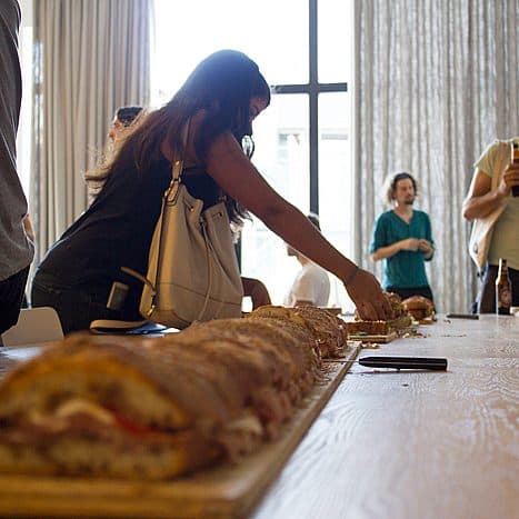 A person is reaching for food from a long sandwich placed on a wooden table. Several other people are standing and chatting in the background. The setting appears to be indoors with natural light coming through large windows.