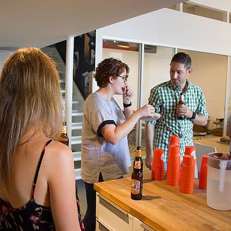 A group of people socializing in an indoor space. A person with short hair and glasses is talking to a person wearing a green checkered shirt. In the foreground, a person with long hair in a floral dress is also present. Red plastic cups and beer bottles are on a table.