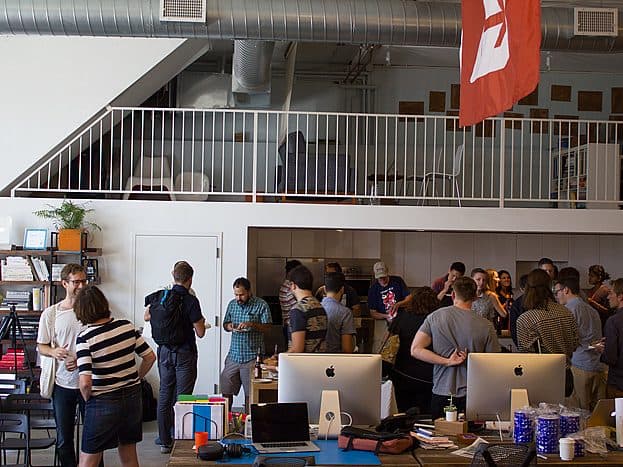 A well-lit, modern office space hosts a social gathering with approximately twenty people, some chatting, some serving themselves food. Multiple desks equipped with iMac computers are in the foreground, and there's a loft and a large red flag in the background.