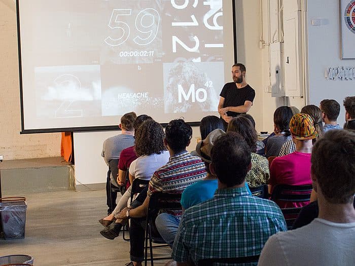 A man stands in front of a room of seated people, giving a presentation. The projector screen behind him displays a countdown timer and various images and numbers. The audience is sitting on black chairs in a minimally decorated room with white walls.
