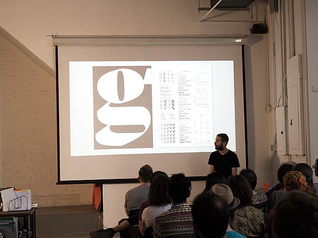 A speaker stands in front of a seated audience, presenting a slide featuring a large stylized letter "g" and various typographic elements on a projector screen. The room appears to be a minimalist industrial space with white walls and exposed pipes.
