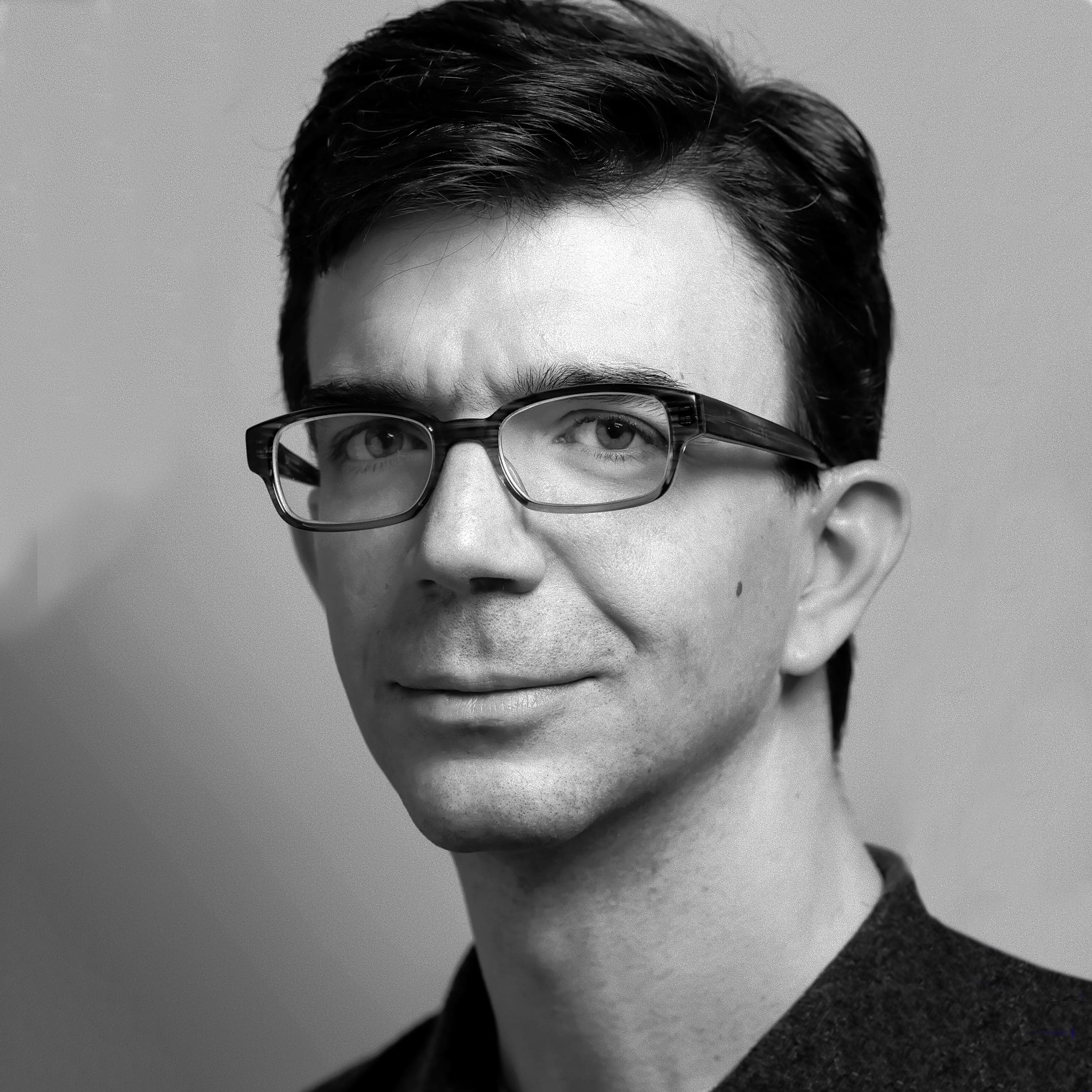 A black-and-white portrait of a man with short dark hair, wearing glasses and a collared shirt. He is looking slightly off to the side with a neutral expression on his face. The background is plain and grayish in tone.