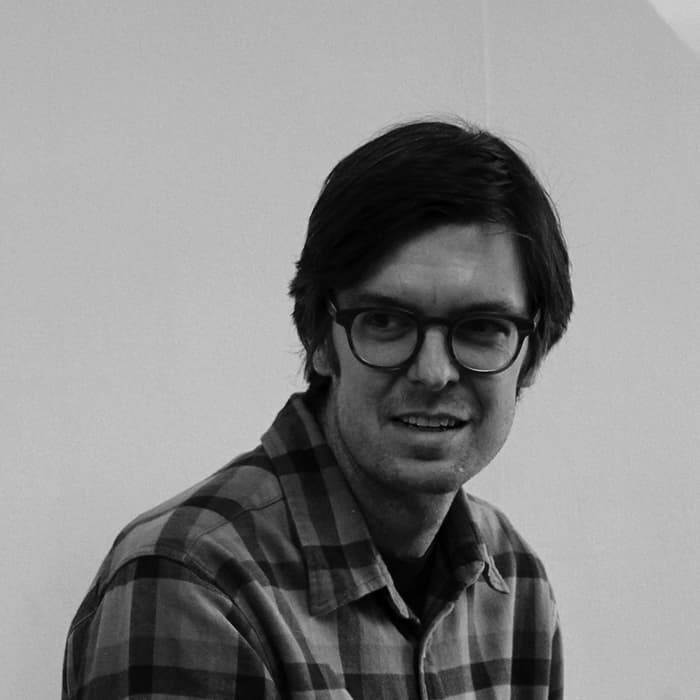 Black and white photo of a person with medium-length dark hair, wearing glasses and a plaid shirt, looking slightly to their left. They are in front of a plain light-colored wall and have a neutral expression.