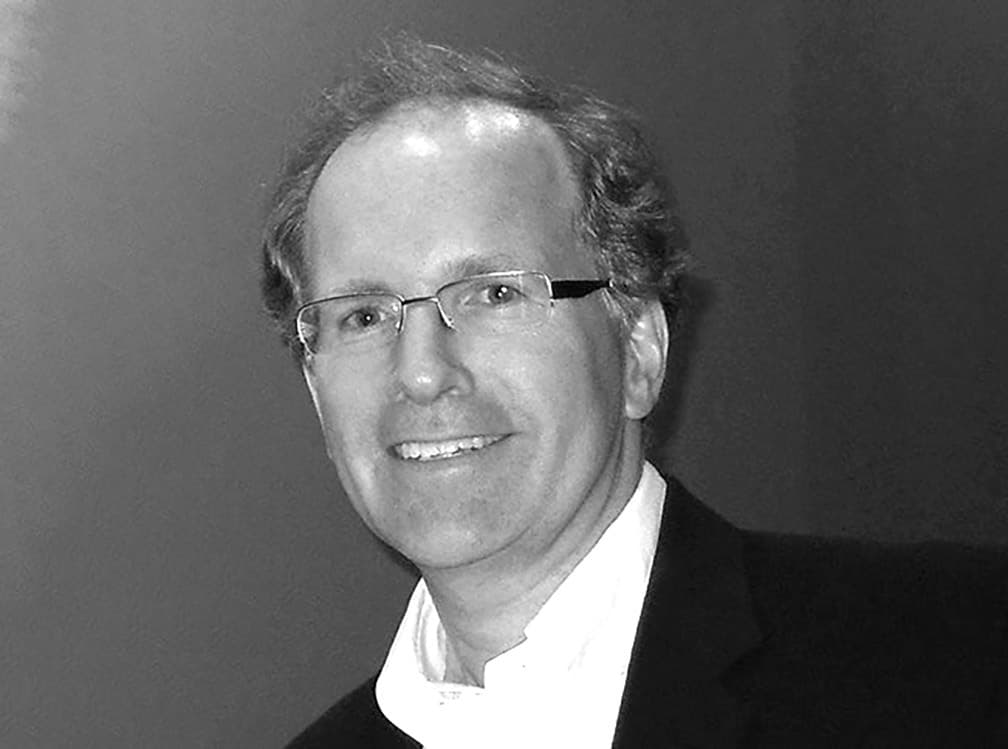 Black and white image of a middle-aged man with curly hair, glasses, and a slight smile. He is wearing a dark suit jacket over a light-colored shirt. The background is plain and neutral.