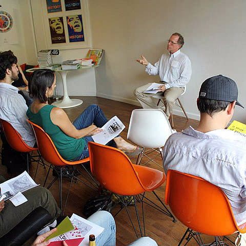A group of people sit in colorful chairs, attentively listening to a person speaking at the front of a room. The speaker, seated on a stool, gestures with their hand while holding a paper. The room has a modern interior with posters on the wall and a plant in the corner.