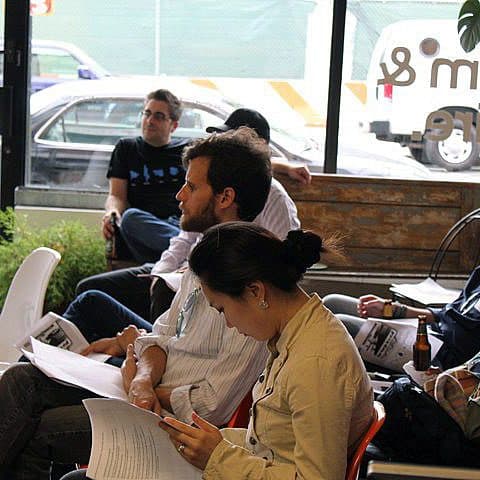 A group of people are sitting indoors, attentively looking at something out of frame. The setting appears to be a casual, modern space with large windows. Some are holding papers or notebooks, while one person is drinking a beverage from a bottle.