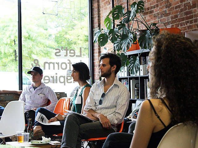 A group of people is sitting in a modern room with large windows and a brick wall. They are facing a speaker, listening attentively. There is a large plant in the background and various chairs and bookshelves in the space. Natural light fills the room.