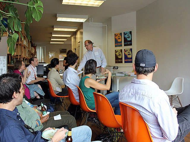 A group of people sit in a room, attentively listening to a man speaking at the front. The room is decorated with colorful wall posters and plants. Some attendees have food and drinks in their hands. The atmosphere appears casual and engaging.