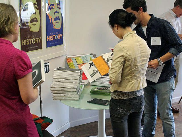 Four people are standing around a table with various books and magazines on it. They are browsing through the materials. The background features posters on the wall. One of the people wears a name tag with "Tim" written on it.