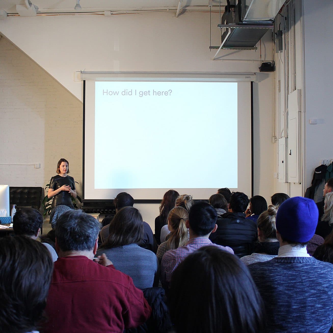 A woman stands in front of a room full of people, giving a presentation. The slide on the screen behind her reads, "How did I get here?" The audience sits attentively, facing the speaker. Coats are hung on wall hooks to the right. The room has a modern, industrial look.