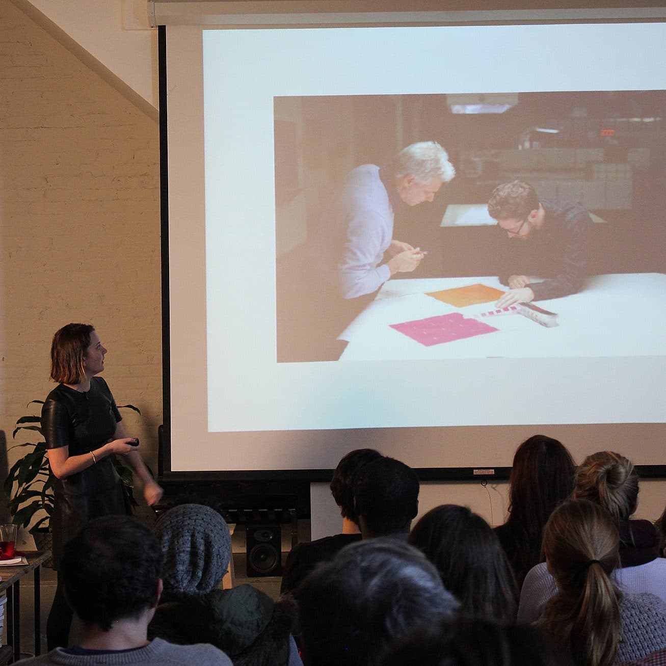 A person stands in front of a seated audience, presenting a slideshow. The current slide shows two individuals examining colorful paper samples. The room has a brick wall, projector screen, and various office items.
