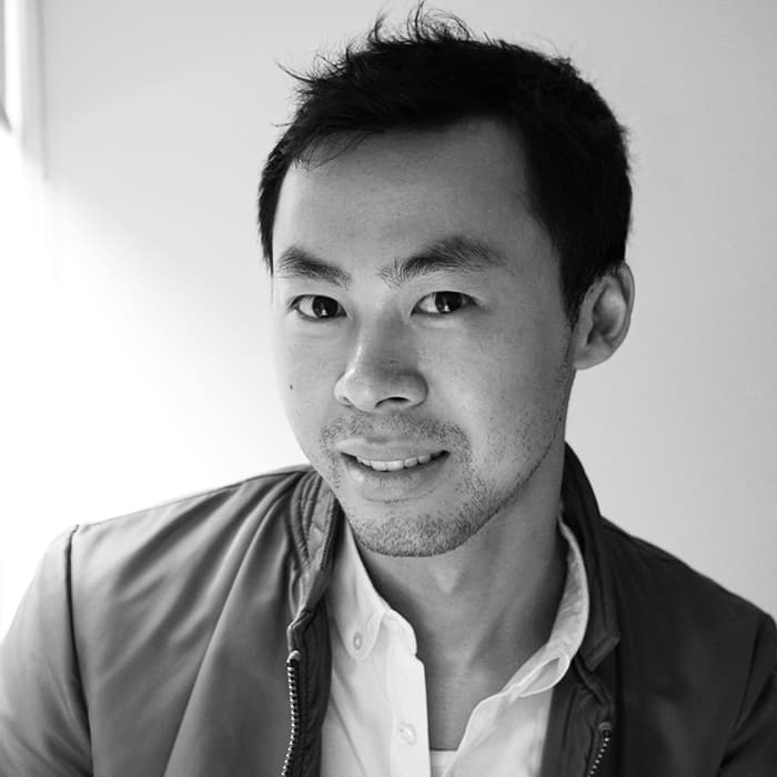 A black-and-white photo of a man with short, dark hair and light stubble. He is wearing a light-colored shirt and a jacket. He is looking slightly off-camera with a subtle smile. The background appears to be a plain, softly lit wall.