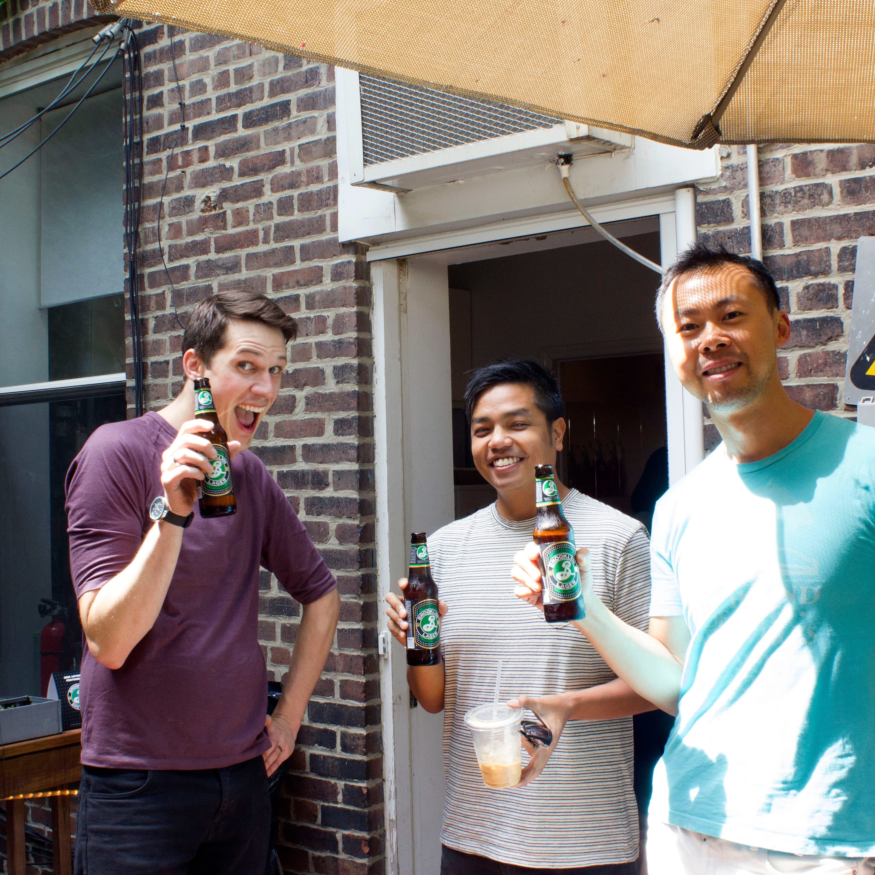 Three people are standing outside under a large umbrella, holding bottles of beer and smiling at the camera. They are near a brick building with a high voltage sign on the wall. One person is holding a cup and the street view includes some plants and electric cables.