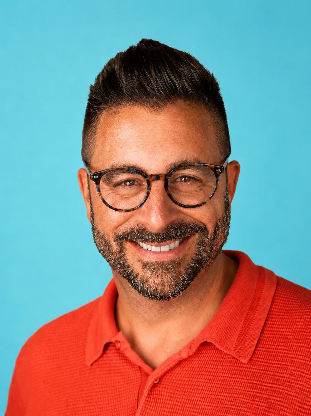 Smiling man with dark hair and beard, wearing round eyeglasses and an orange collared shirt, standing in front of a plain light blue background.