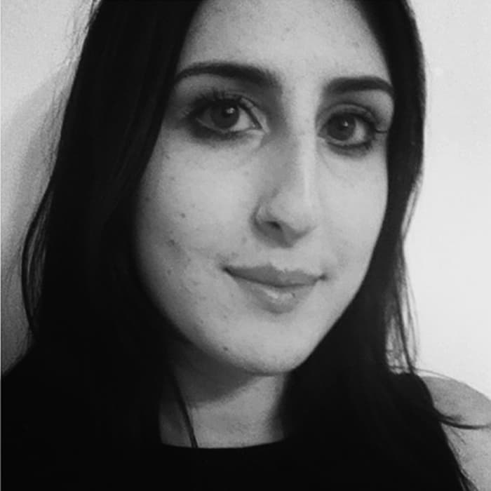 Black and white close-up portrait of a person with long dark hair and dark eyes, slightly smiling, looking straight at the camera. The background is plain, suggesting they are indoors. The lighting is soft, casting subtle shadows on the face.