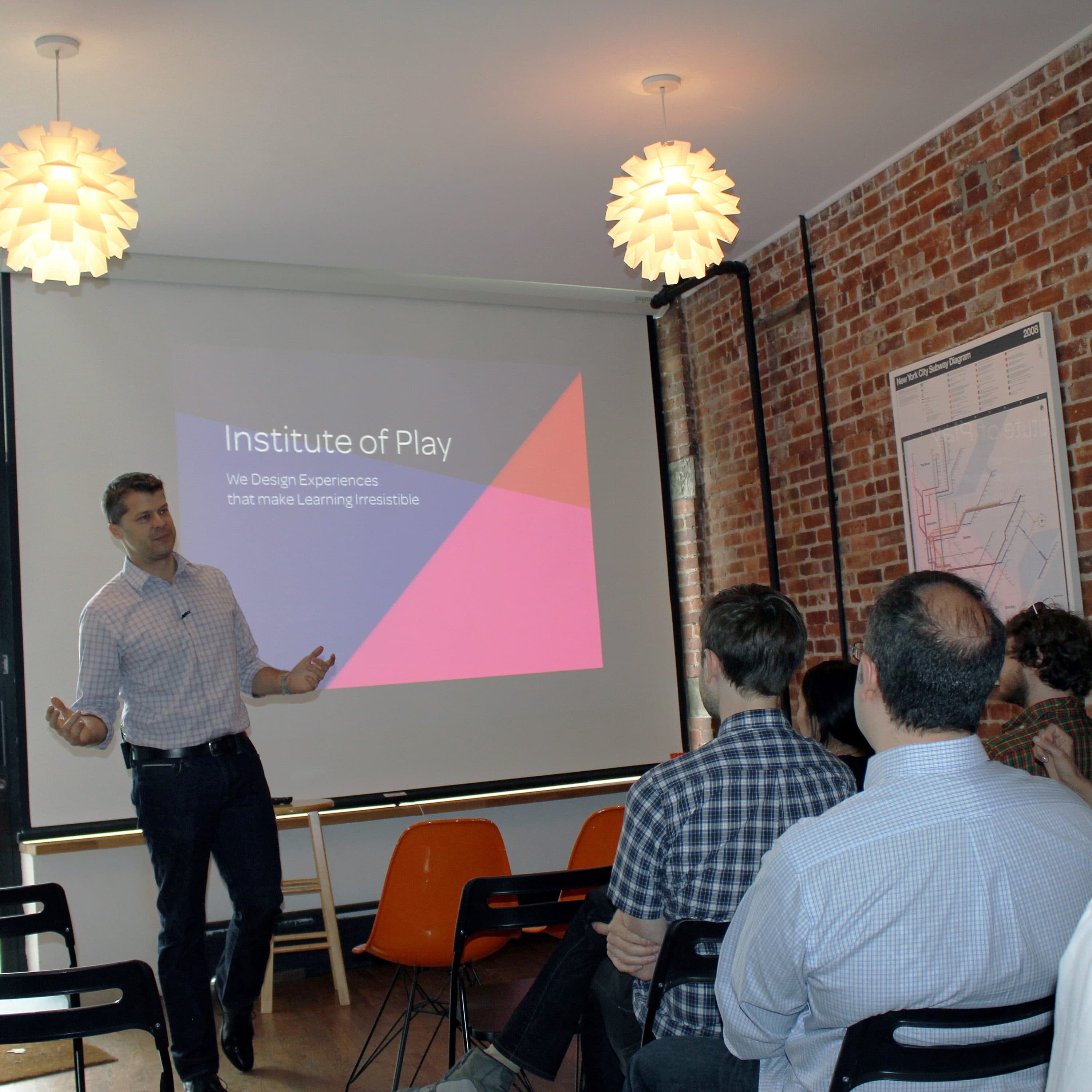 A man and woman present to a seated audience in a room with brick walls. A projector screen behind them displays the text "Institute of Play: We Design Experiences That Make Learning Irresistible." The room is well-lit with two hanging lamps.
